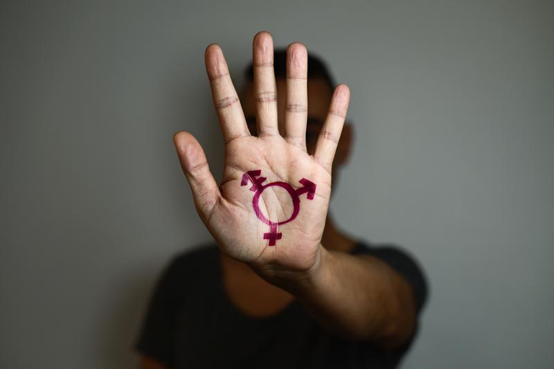 a light skilled person raising their hand towards the camera with a purple symbol for trans people painted on their hand